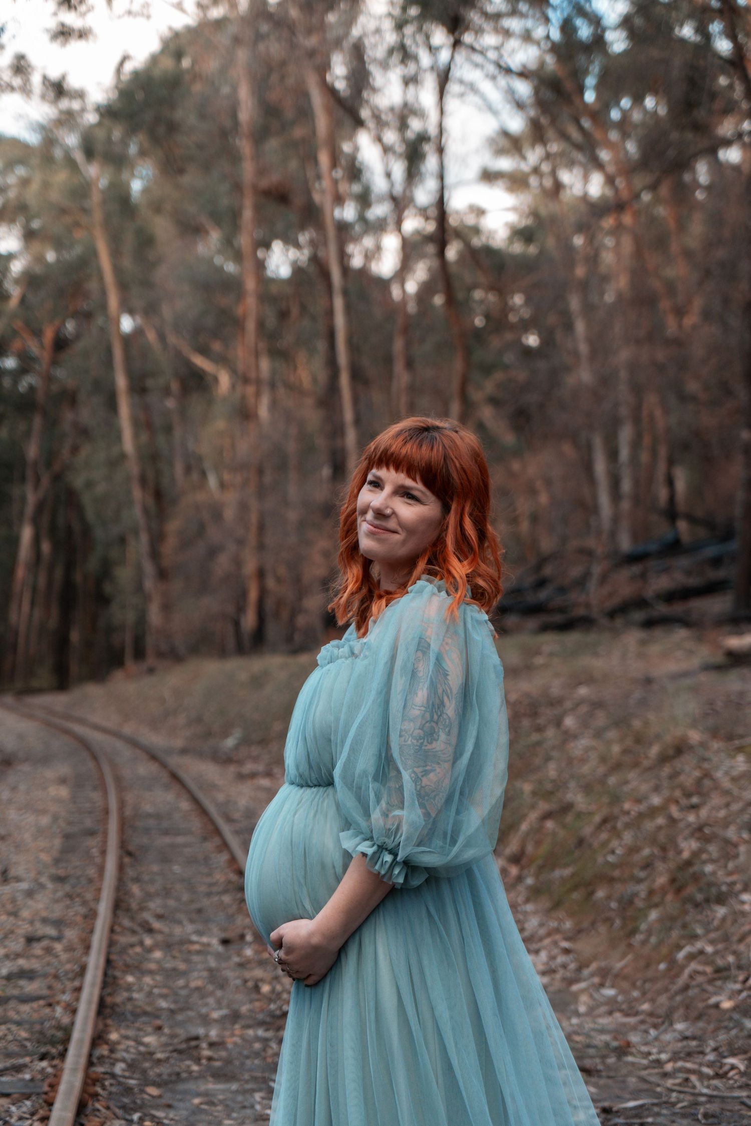 Pregnant woman in blue dress standing by railroad tracks in a forest setting.