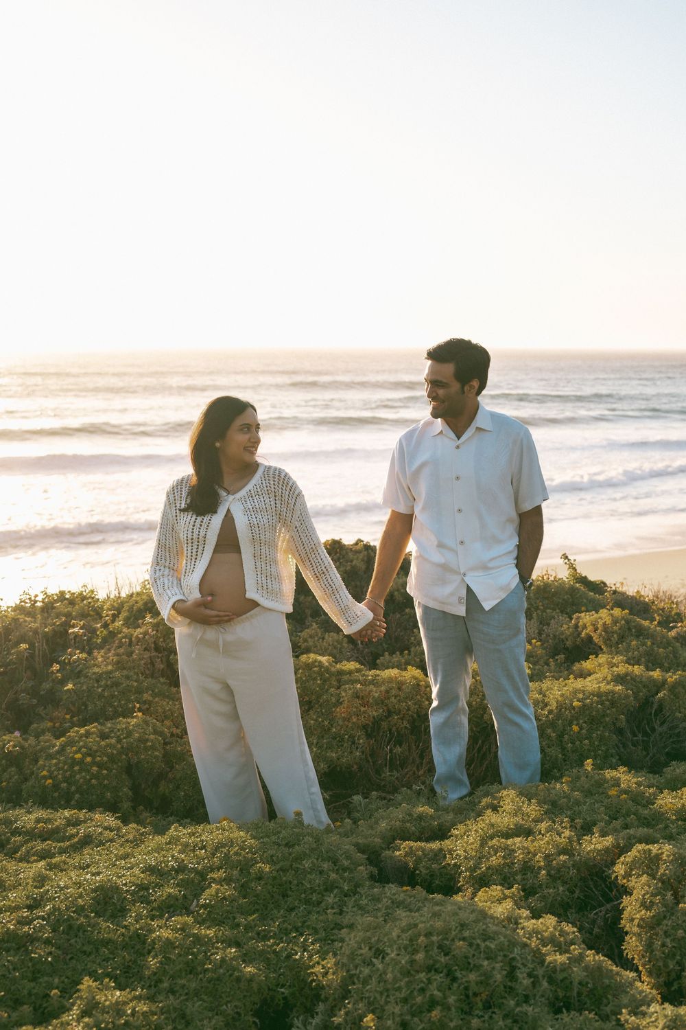 Pregnant couple holding hands on a beach at sunset, woman cradling baby bump, ocean waves in background.