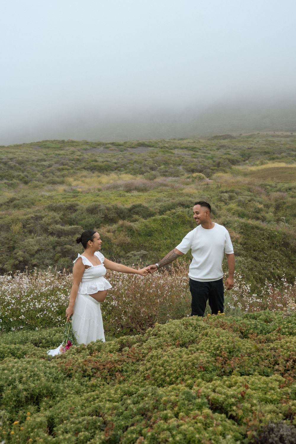 Couple holding hands in a misty coastal meadow, woman in white dress and man in white shirt smiling at each other.