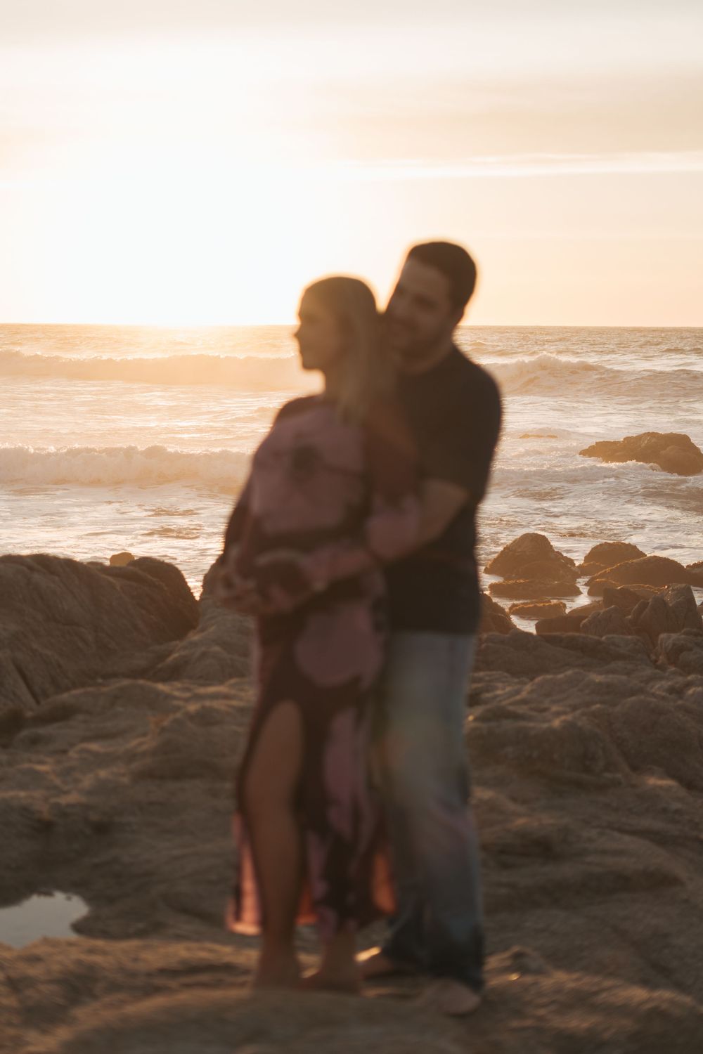 A couple stands together on rocky coastal cliffs at sunset while waves crash against the shore in the background.