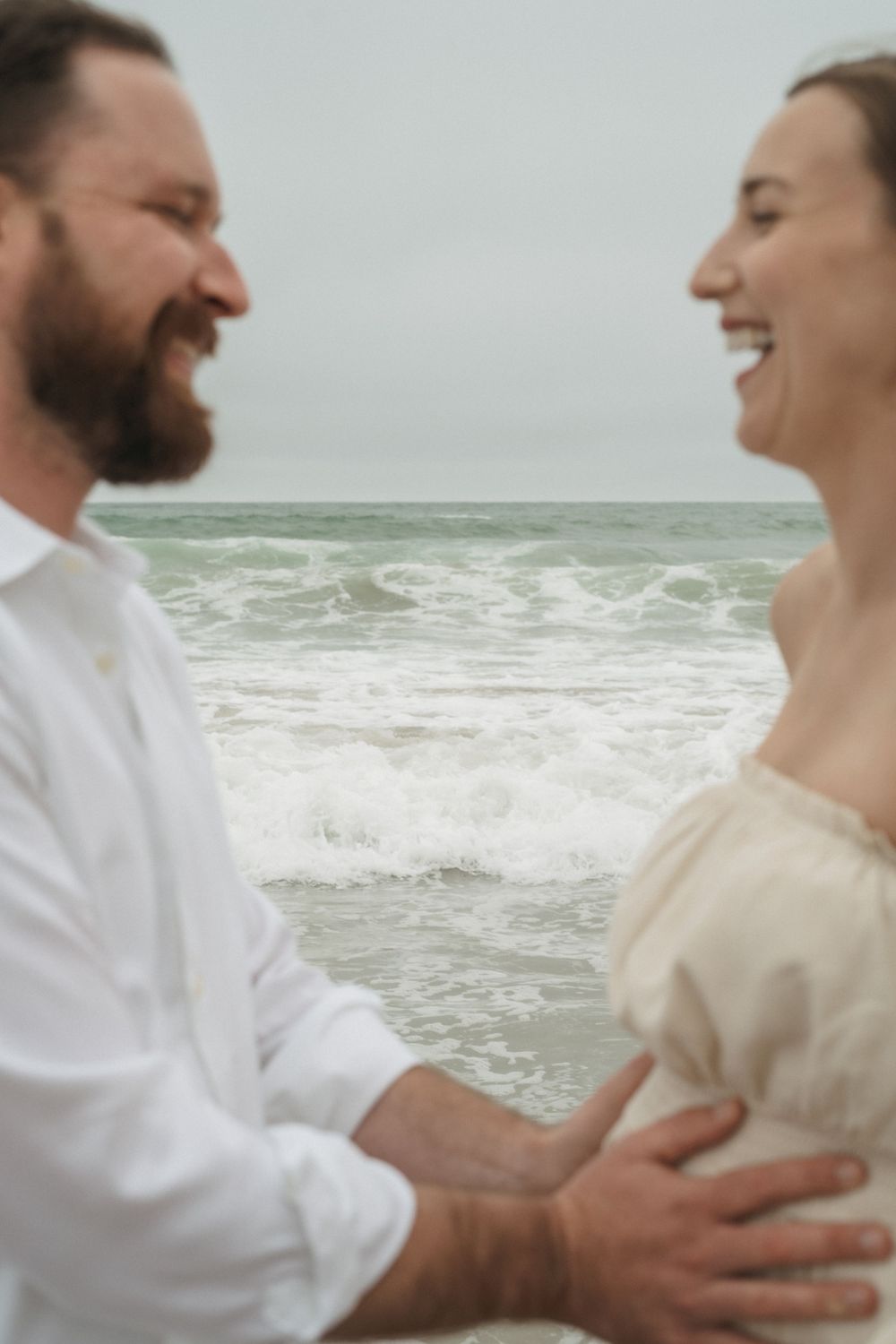 A joyful couple shares a laugh together on a stormy beach while wearing light-colored clothing.