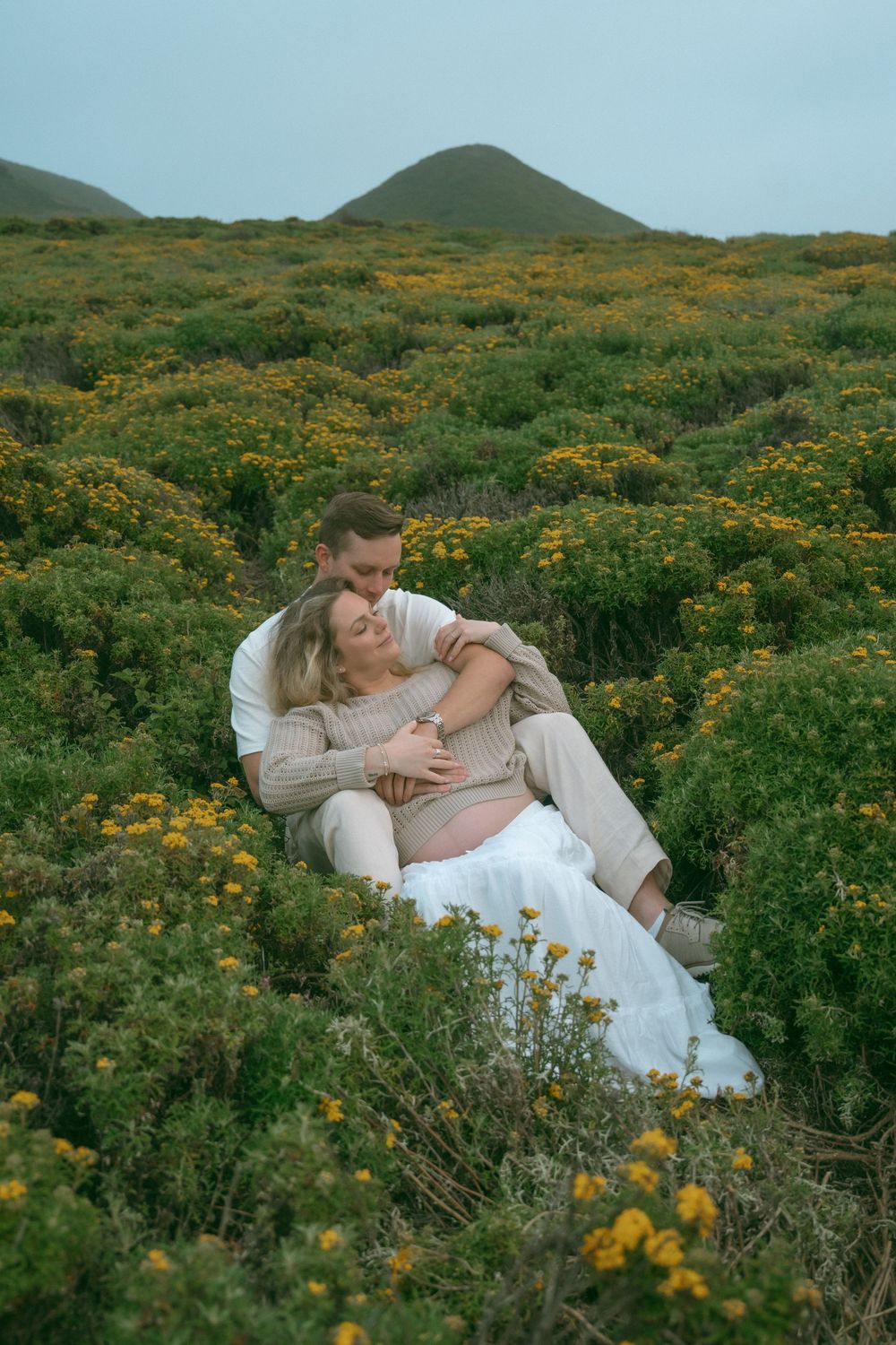 Couple embracing in a field of yellow wildflowers with green hills in the background.
