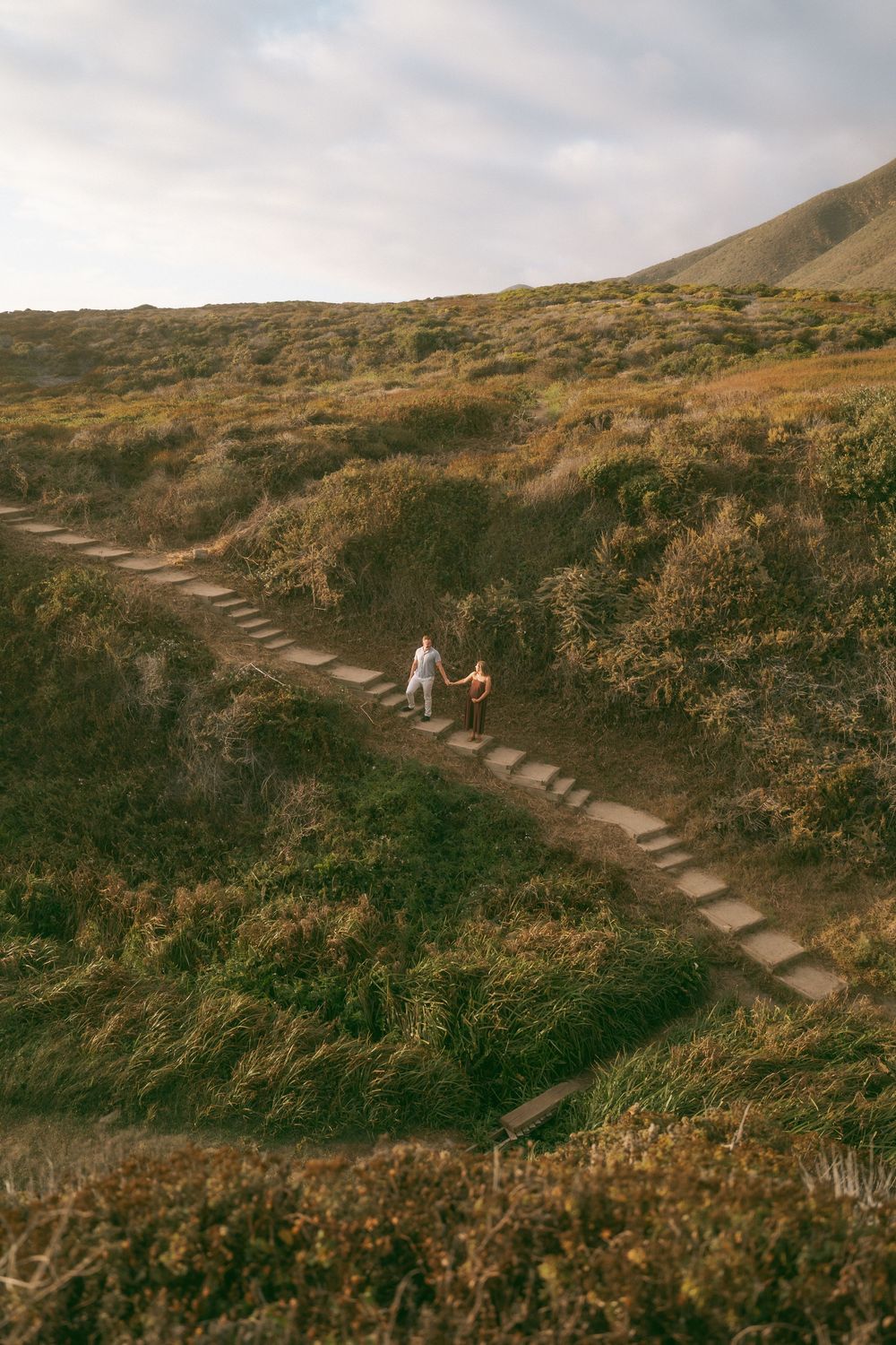 Two hikers walk along a winding stone path through lush green hillside vegetation under a cloudy sky.
