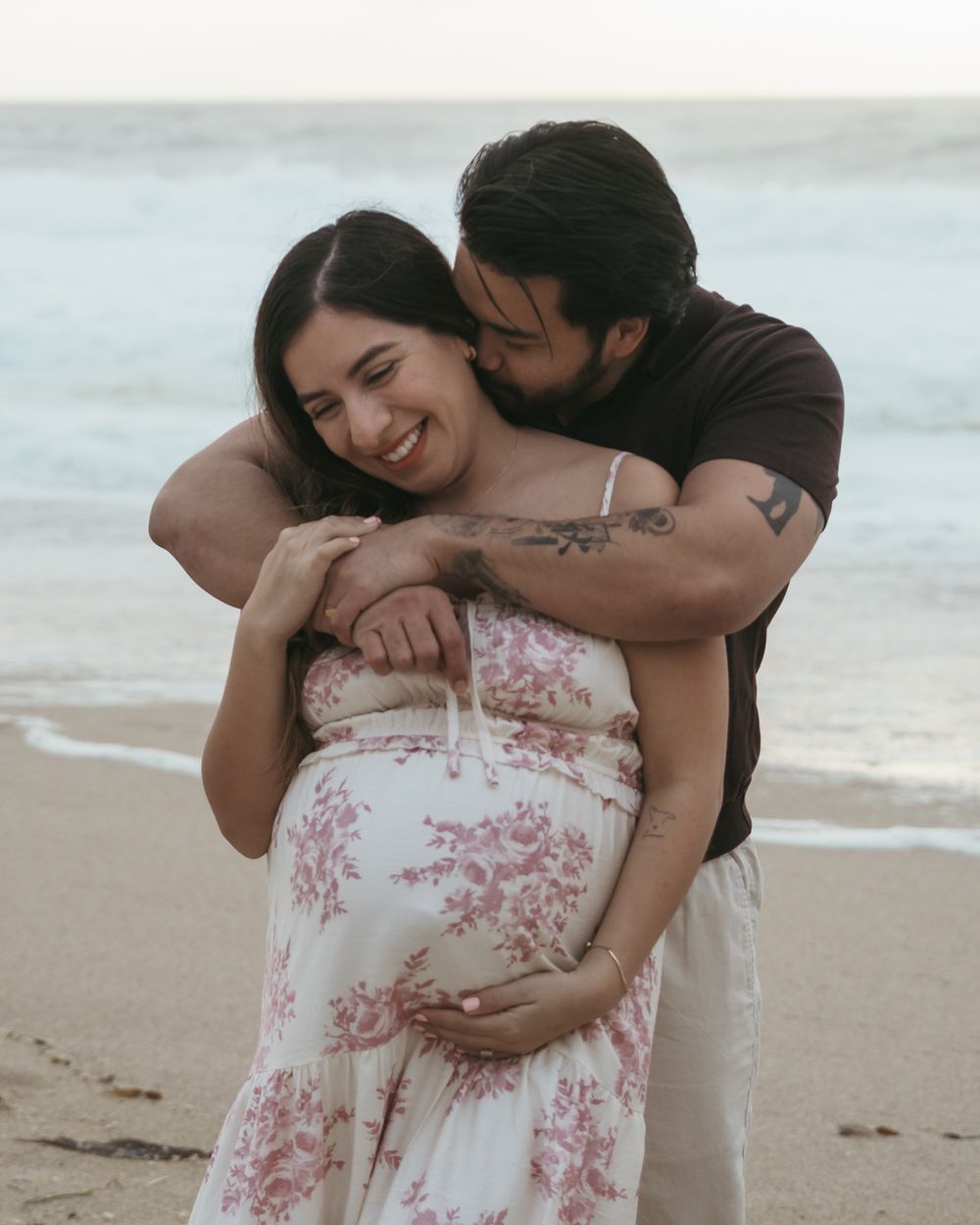 A couple shares an intimate moment on a beach at sunset while wearing a floral dress and dark clothing.