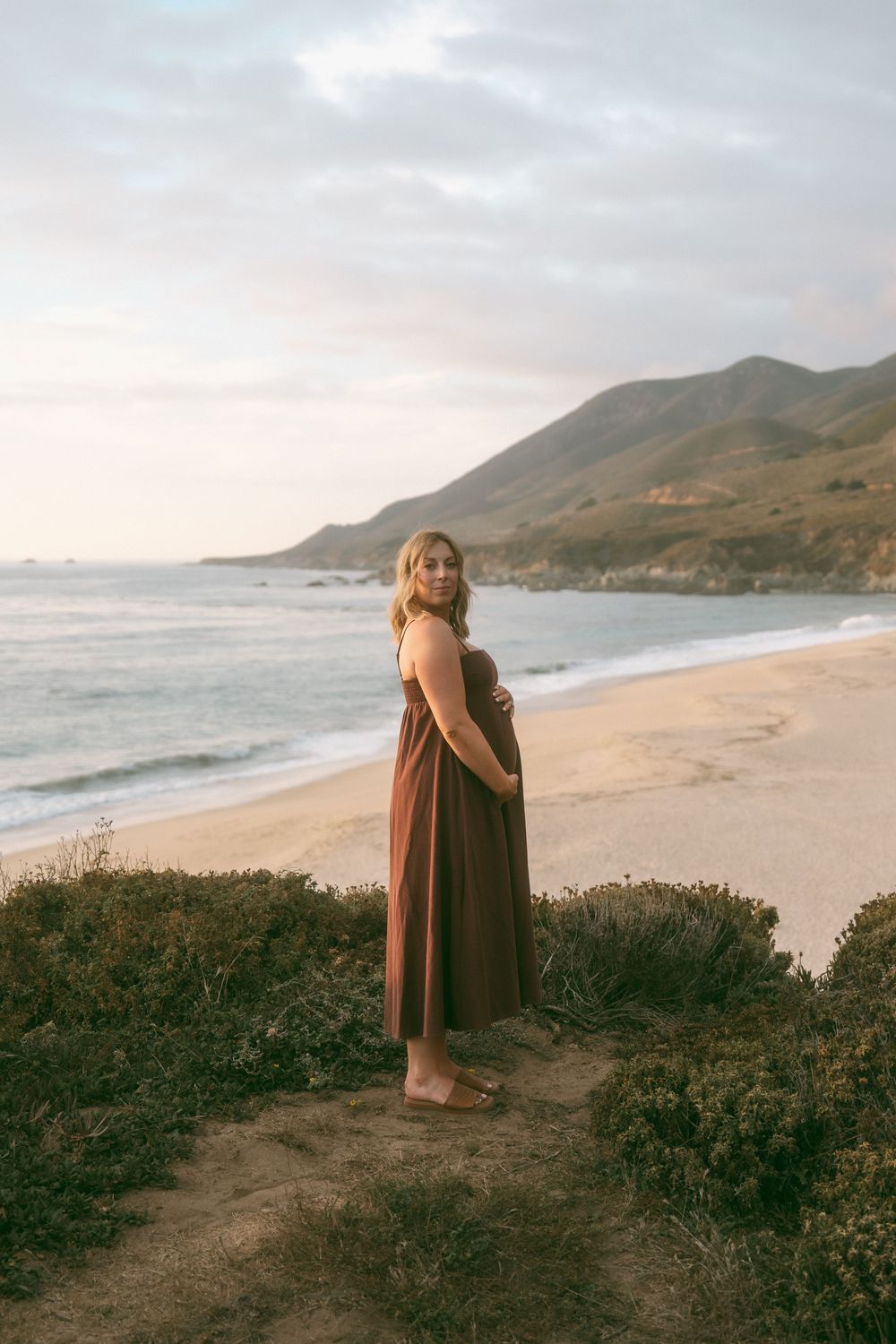 Pregnant woman in brown dress standing on coastal bluff at sunset, cradling baby bump with ocean and mountains behind.