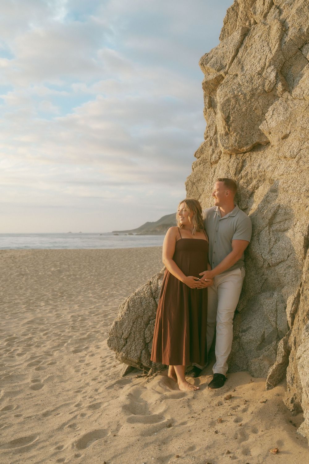 Couple leaning against a large rock on a sandy beach at sunset, woman in brown dress, man in gray polo.