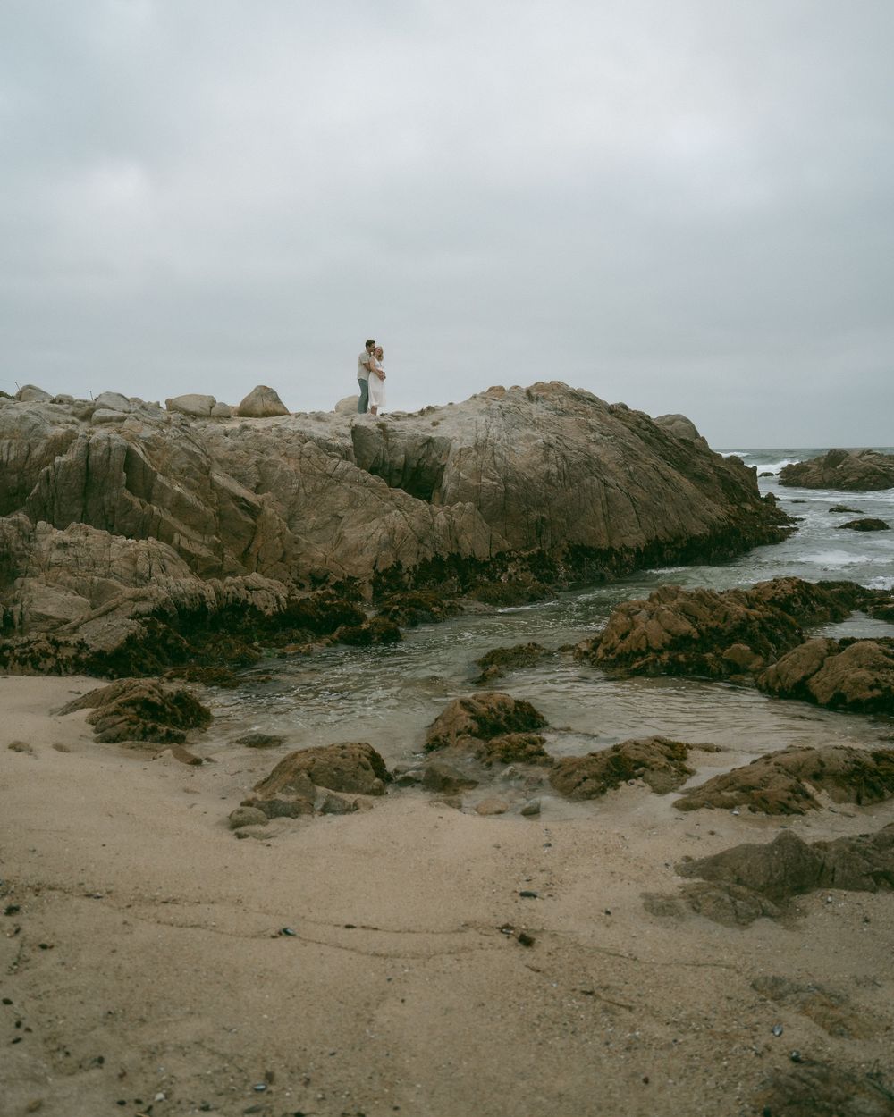 Couple standing on rocky coastal cliffs embracing, with sandy beach and calm ocean waters below on an overcast day.