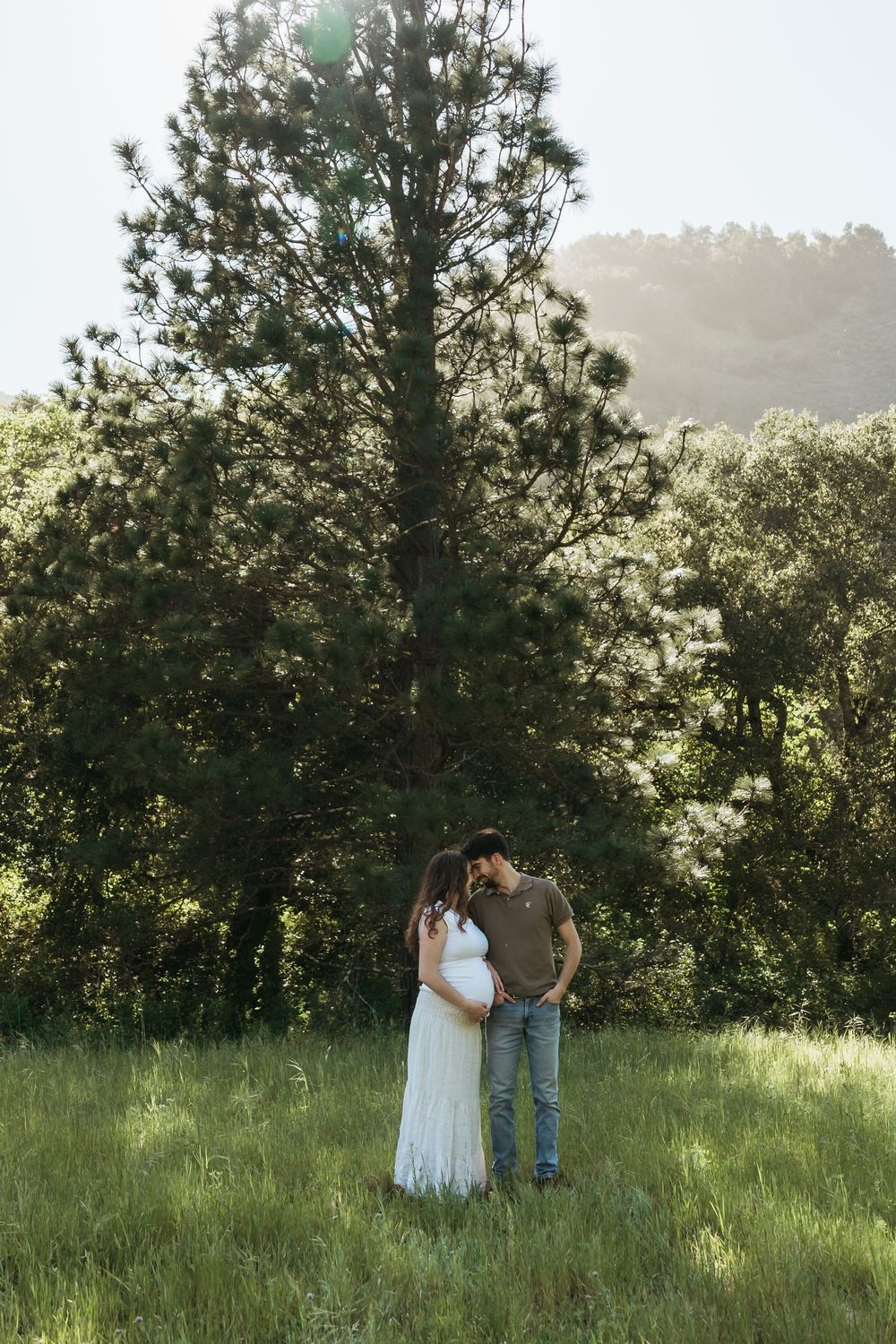 A couple shares an intimate moment in a sunlit meadow surrounded by trees during a golden hour photoshoot.