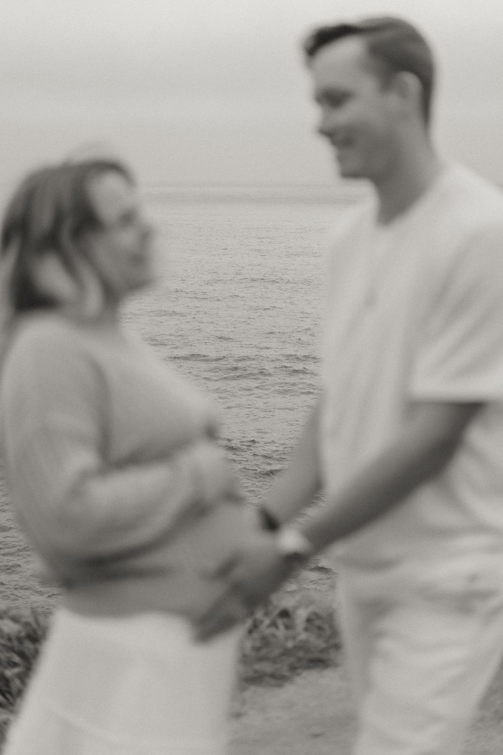 Black and white photo of a couple holding hands by the ocean, woman visibly pregnant, sharing a tender moment.