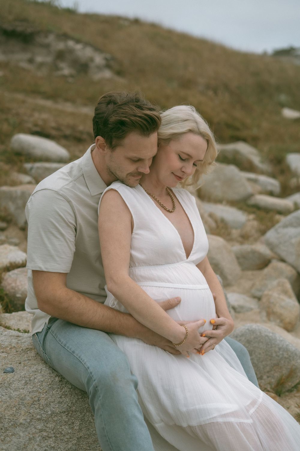 Couple sitting on rocks outdoors, man embracing pregnant woman in white dress, sharing a tender moment in nature.