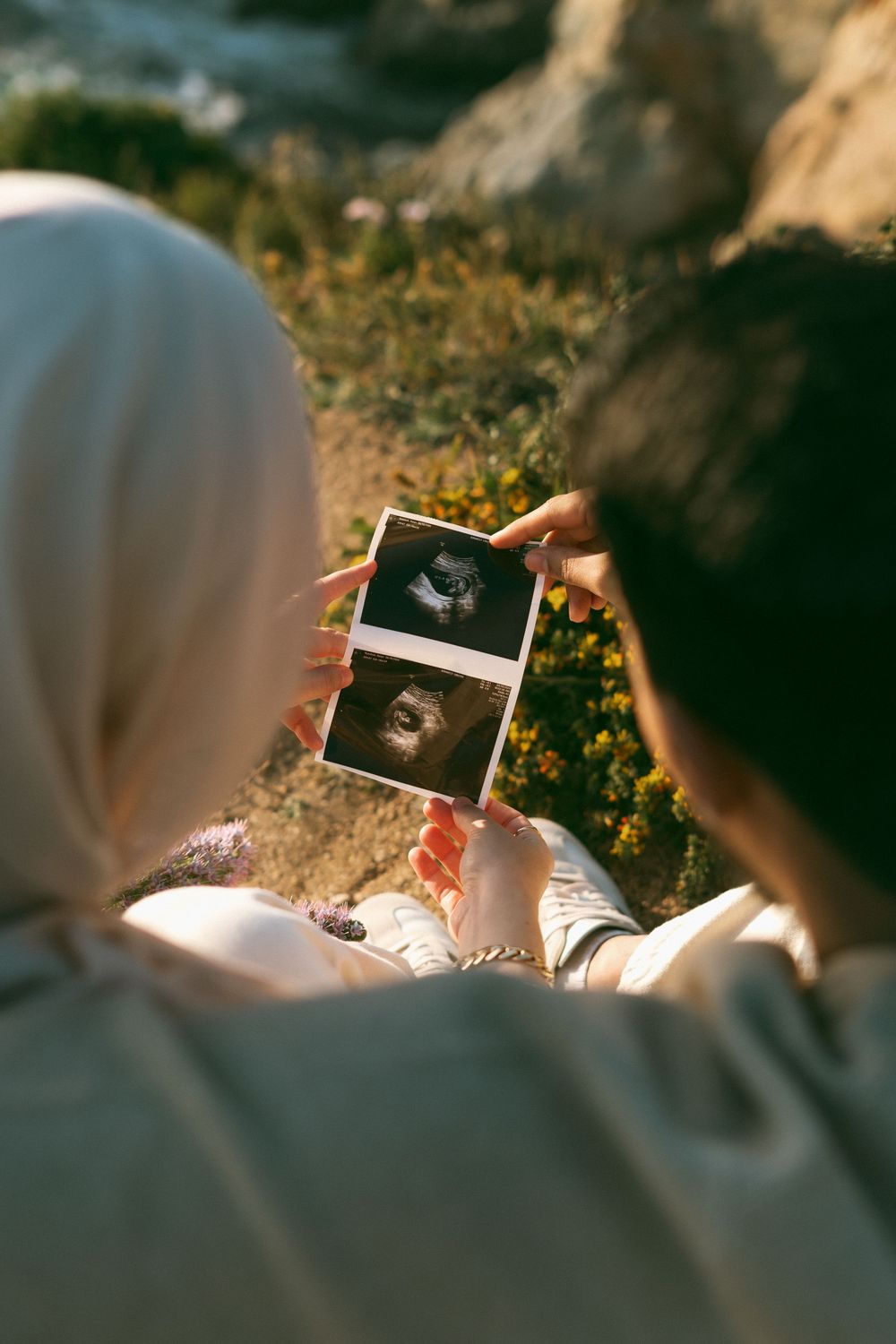 Two women, one wearing a hijab, looking at ultrasound photos together outdoors in natural sunlight.