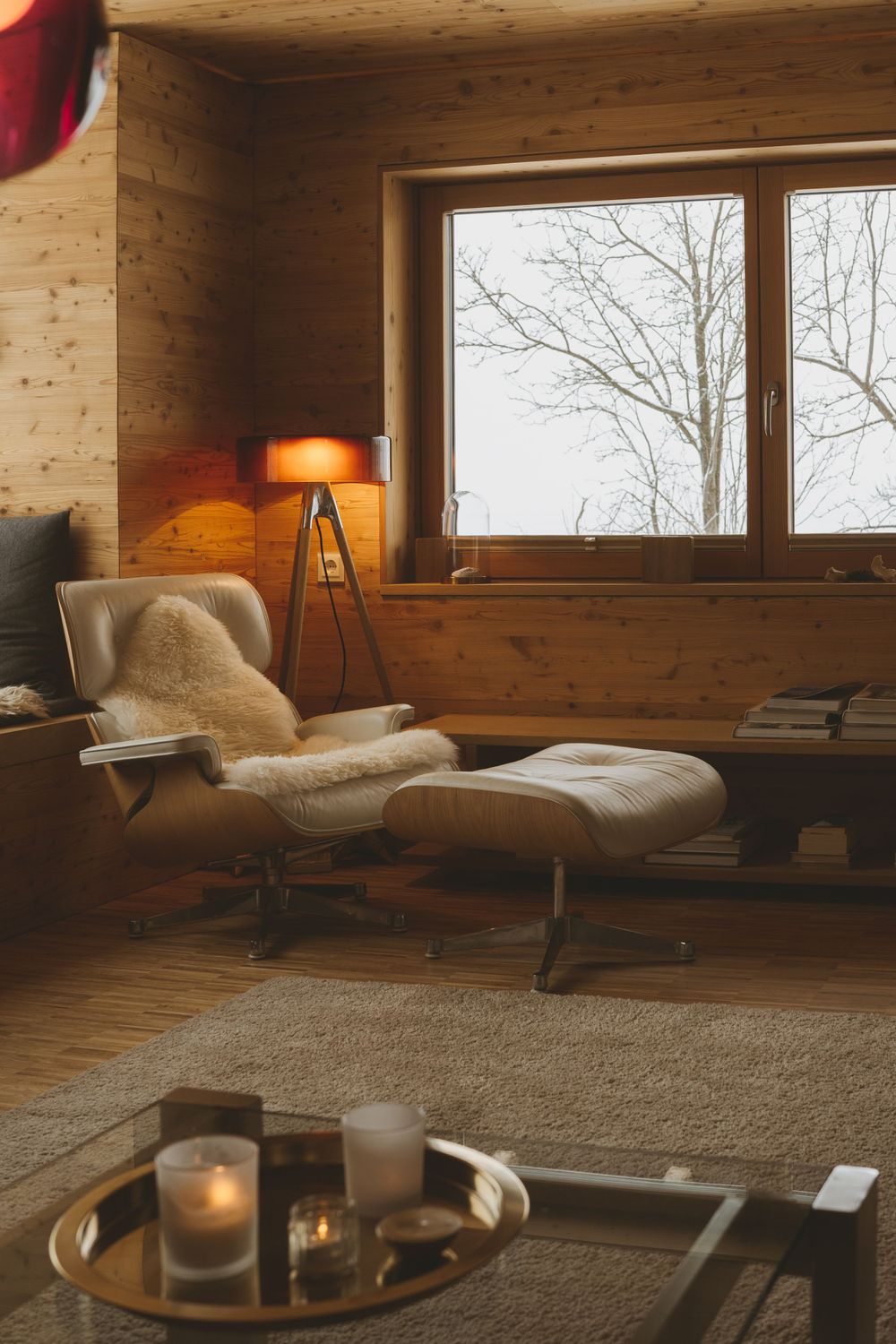 Cozy modern cabin interior with leather lounge chair, fur throw, and warm lighting beside window.