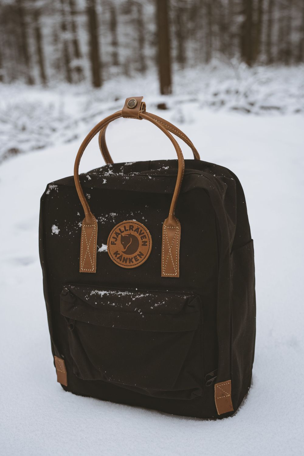 Black Fjallraven Kanken backpack with brown leather straps sitting in snow-covered winter forest.