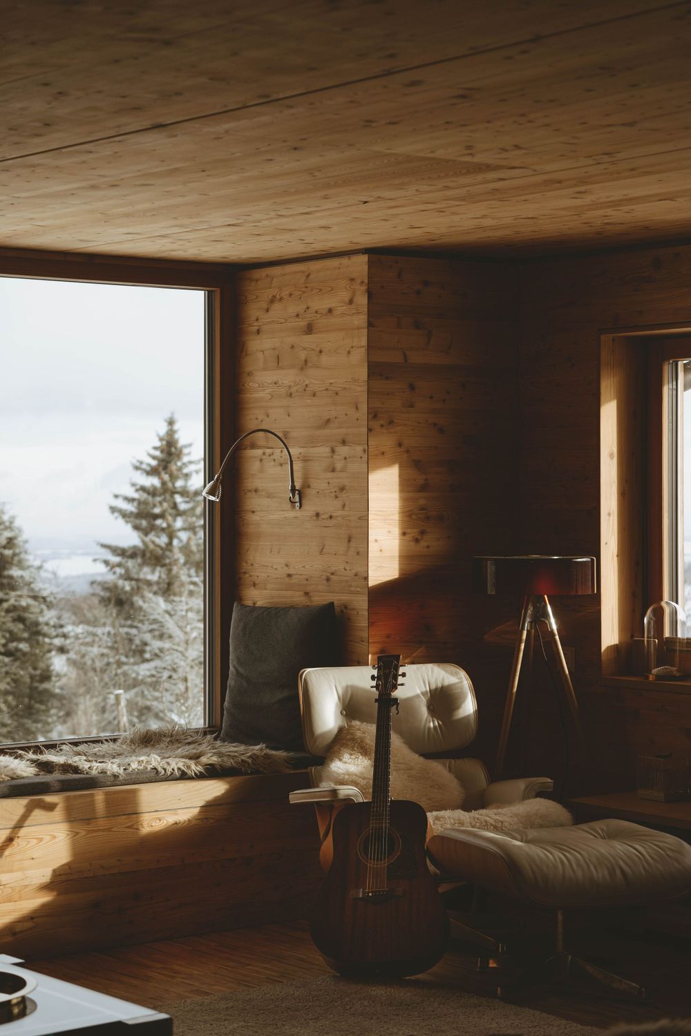 Modern cabin interior with large windows, wooden walls, and a cello by the window overlooking snowy landscape.