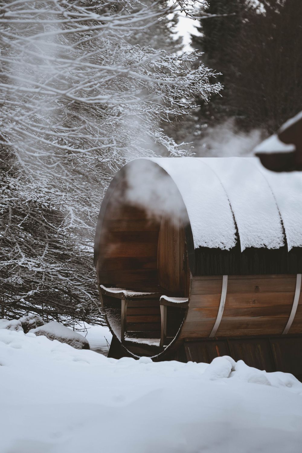 Snow-covered wooden barrel sauna nestled among frost-covered trees in winter.