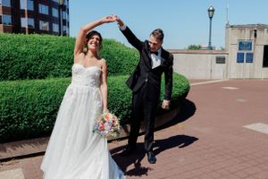 A couple celebrates during their wedding day dance on a sunny brick patio with greenery and buildings in the background.