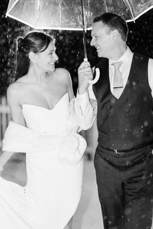 Black and white photo of a bride and groom smiling under a clear umbrella in the rain on their wedding day.
