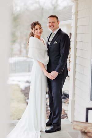 Bride and groom pose together on a porch in winter, she in a white gown with wrap, he in a dark suit.