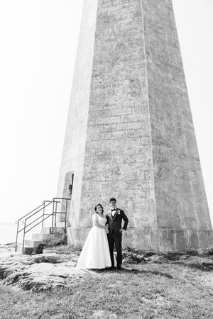Black and white photo of bride and groom standing together at base of tall lighthouse on rocky shoreline.