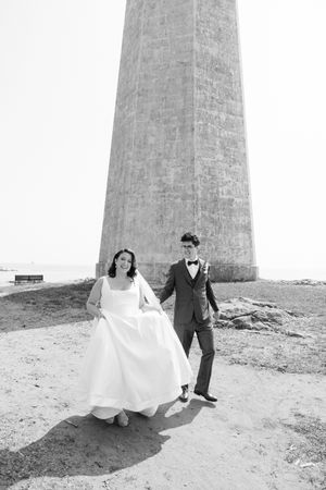 Black and white photo of bride and groom walking hand in hand in front of a tall concrete lighthouse.