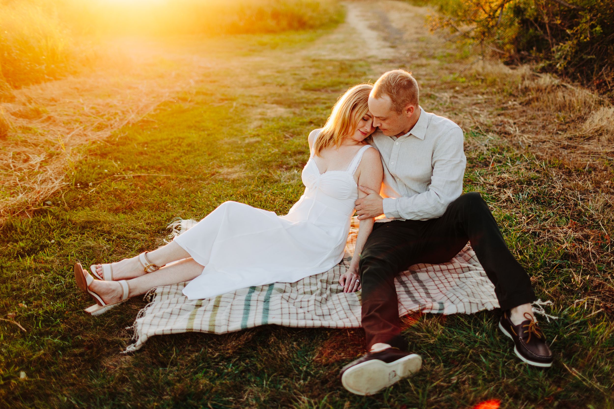Couple sitting on a blanket in a sunlit field, sharing a tender moment during a golden hour engagement photo session.