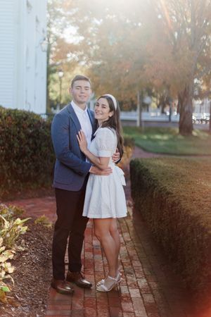 Couple embracing outdoors on brick path, man in blue blazer, woman in white dress, surrounded by hedges and autumn trees.