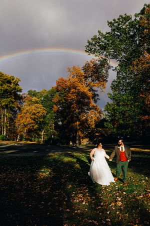 Bride and groom walk hand-in-hand through autumn park with vibrant rainbow arching over colorful fall trees.