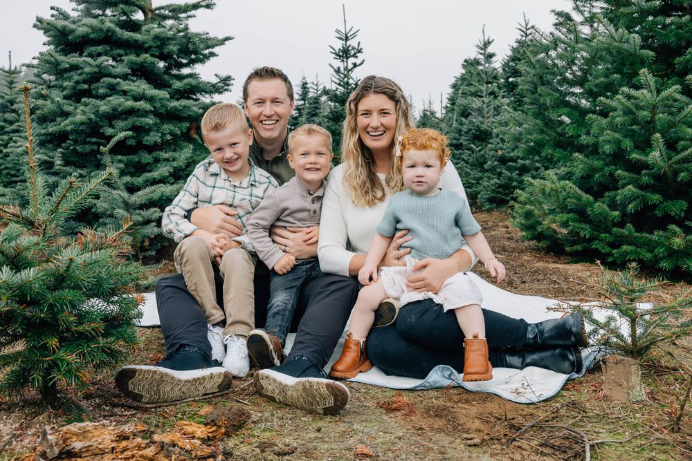Happy family of five sitting on a blanket surrounded by lush green Christmas trees at a tree farm.