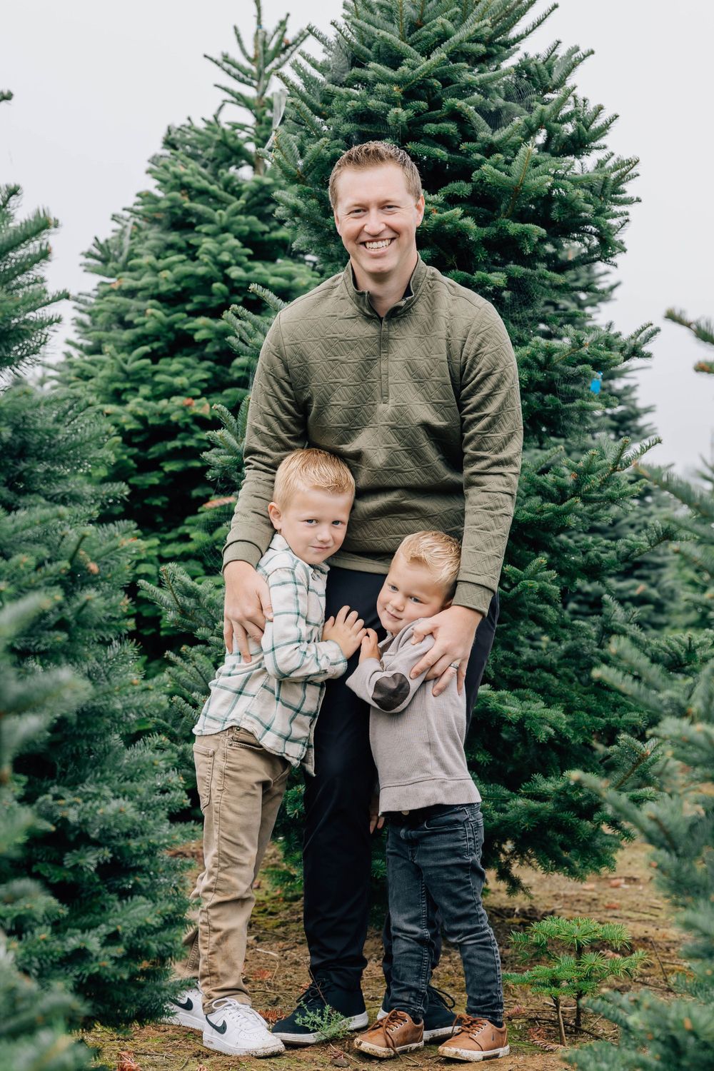 Tall father smiling with his two young sons in front of lush Christmas trees outdoors.