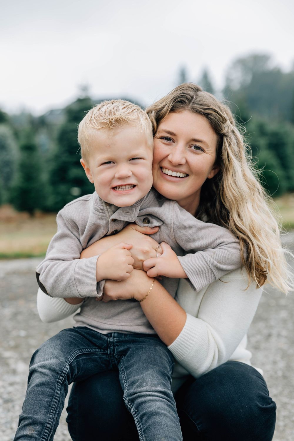 Loving mother hugging her young blonde son while both smile outdoors at a tree farm.