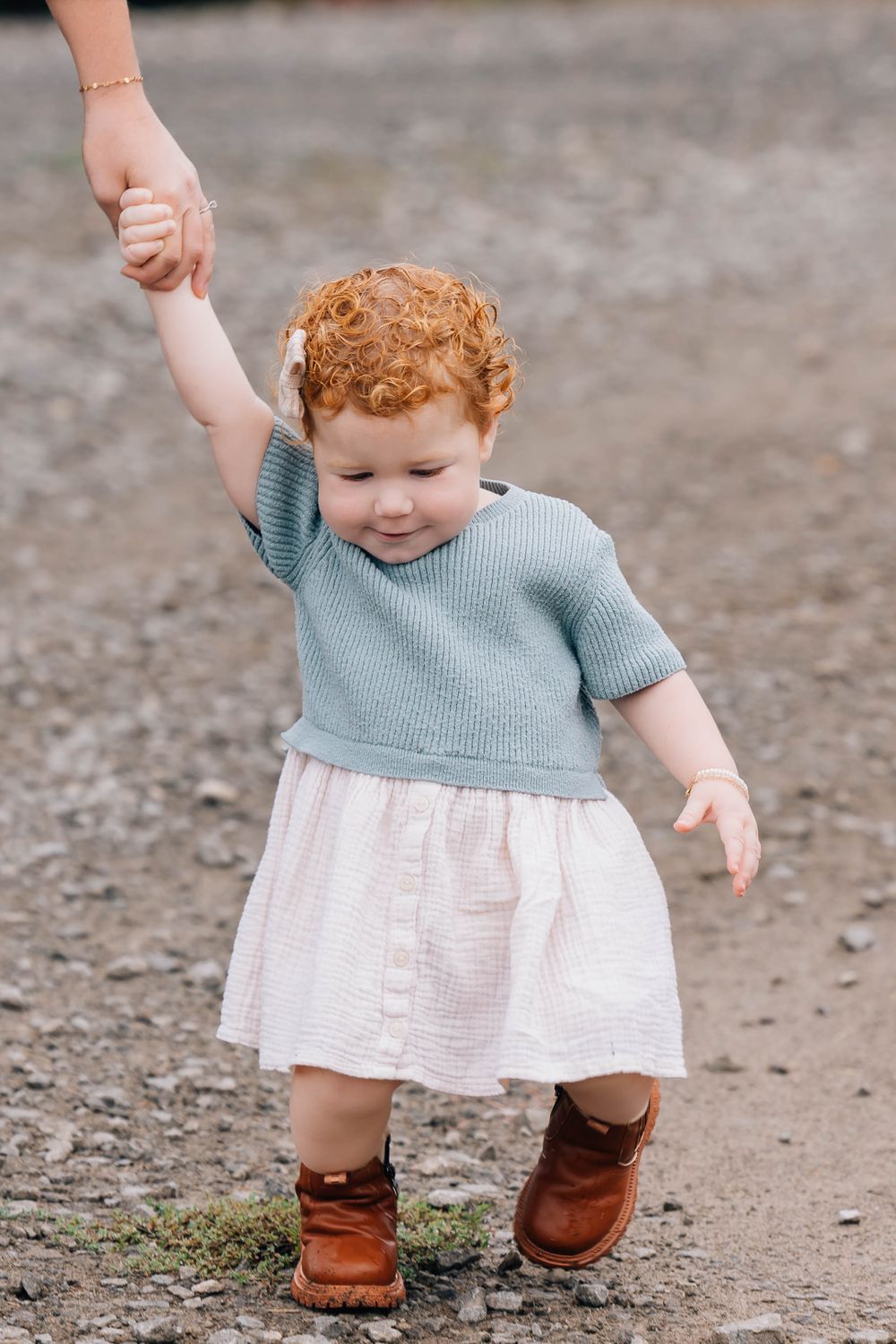 Adorable red-haired toddler girl in a teal sweater and skirt holding an adult hand outdoors.