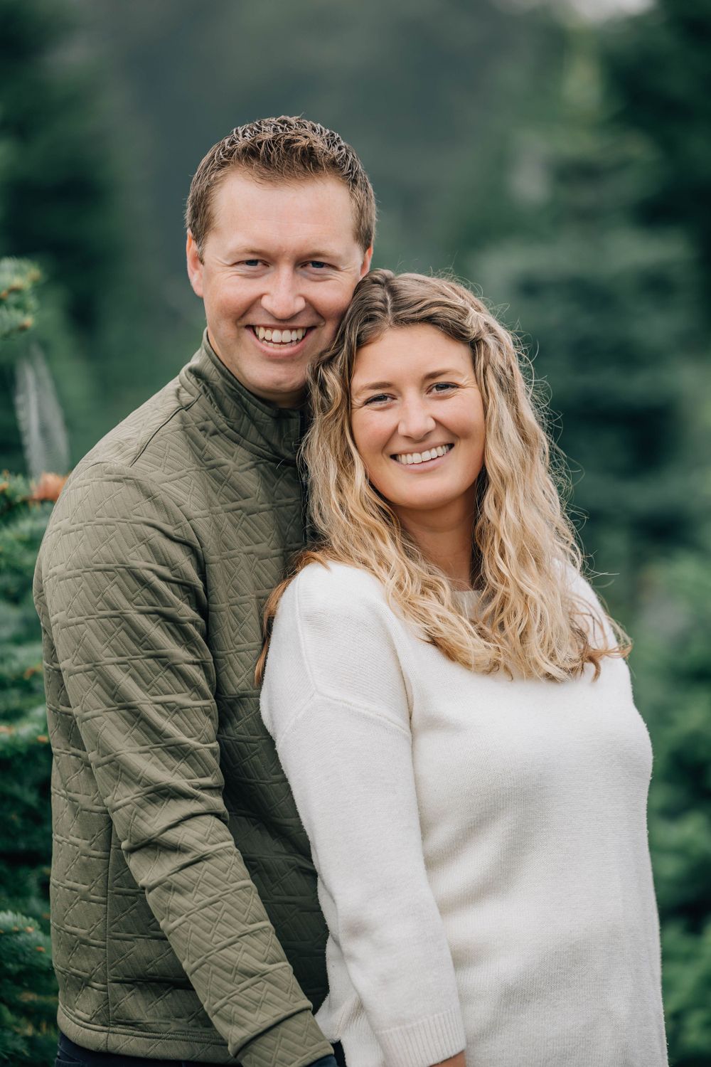 Smiling couple posing together outdoors surrounded by green trees in a warm embrace.