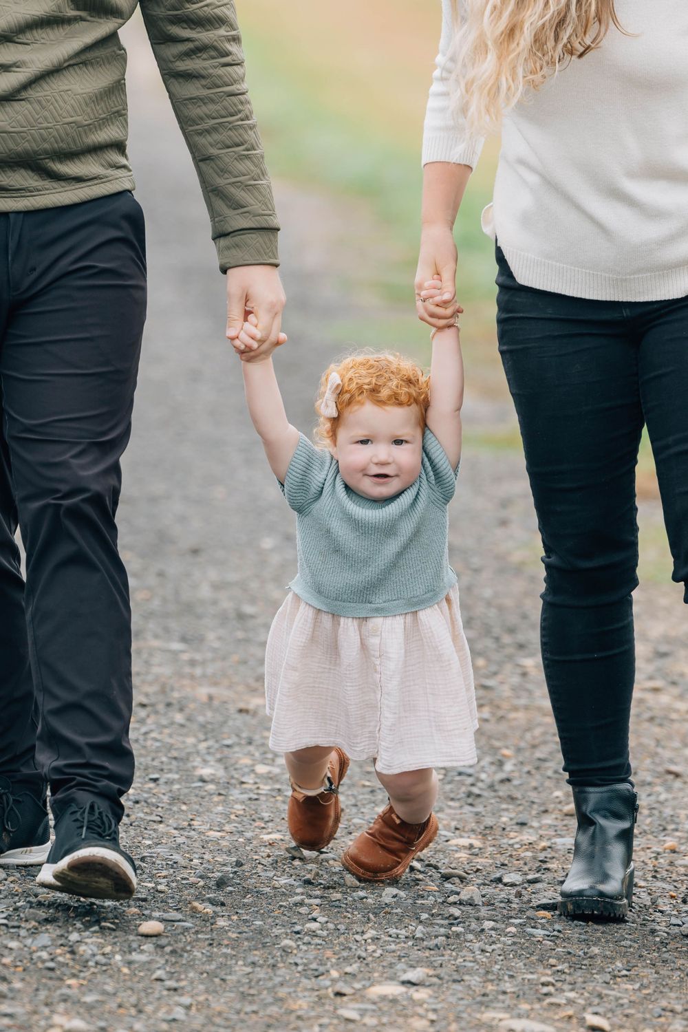 Adorable red-haired toddler girl in a blue sweater and skirt being lifted by parents hands on a gravel path.