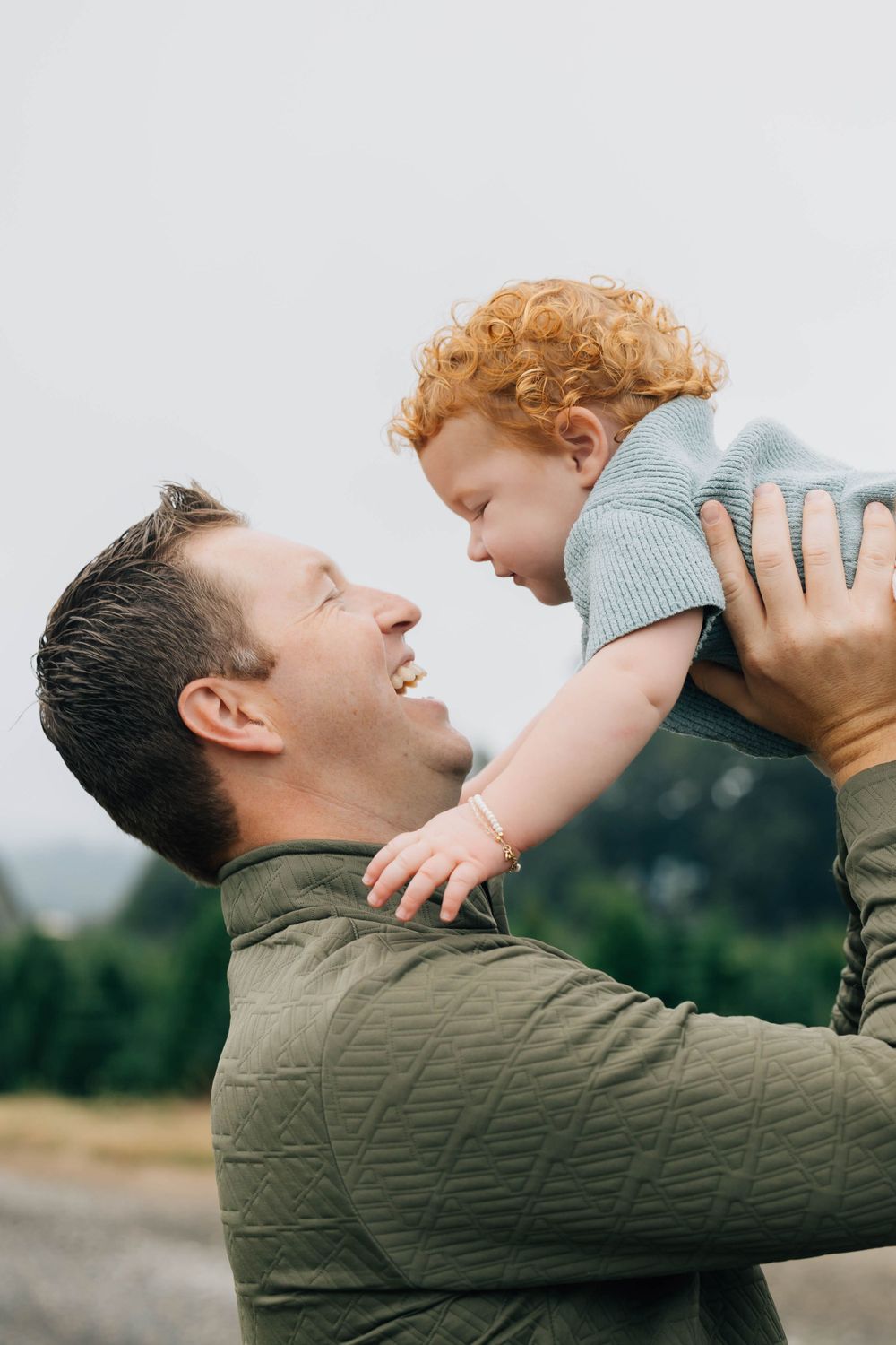 Laughing father lifting his joyful red-haired baby girl up in the air outdoors.