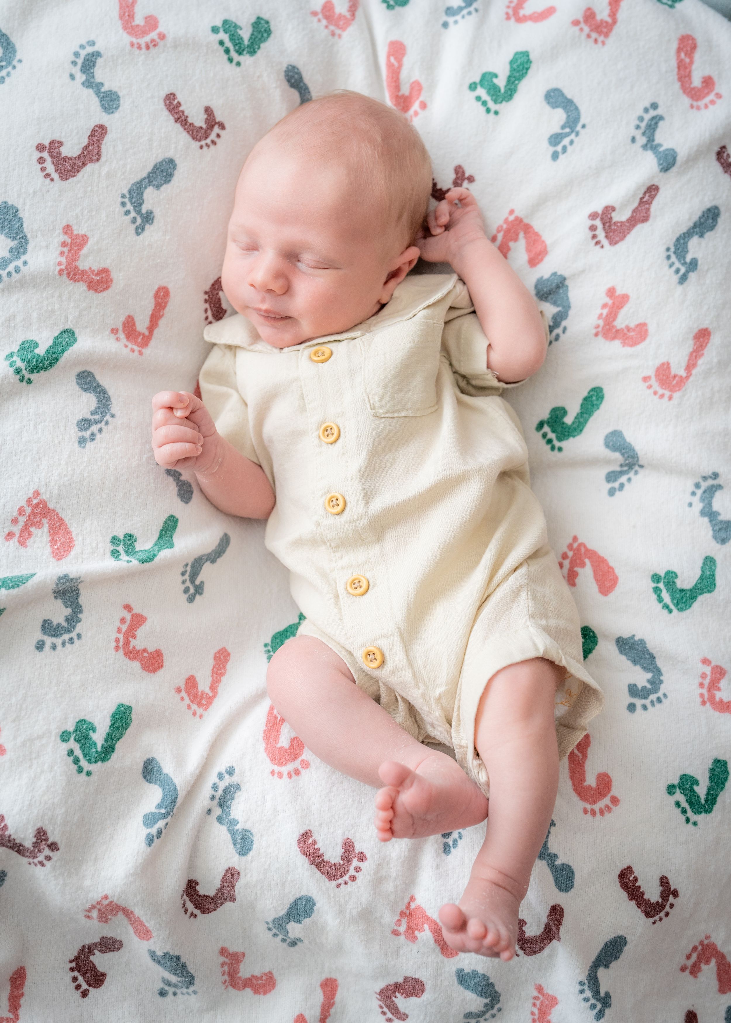 Sleeping newborn infant wearing cream-colored clothing on a patterned blanket with footprint designs.