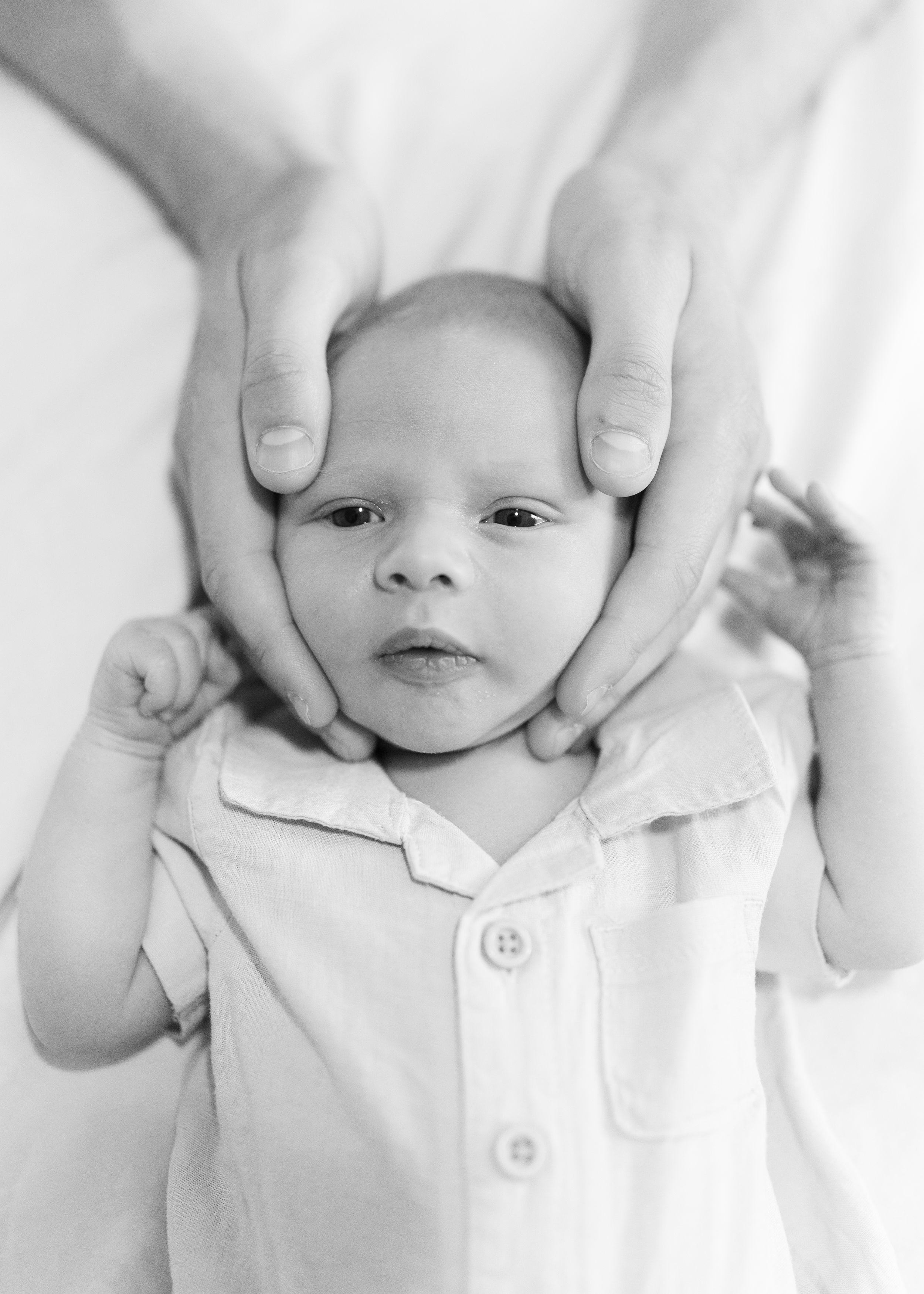 Black and white portrait of a newborn baby in white clothing with hands gently cradling their head.