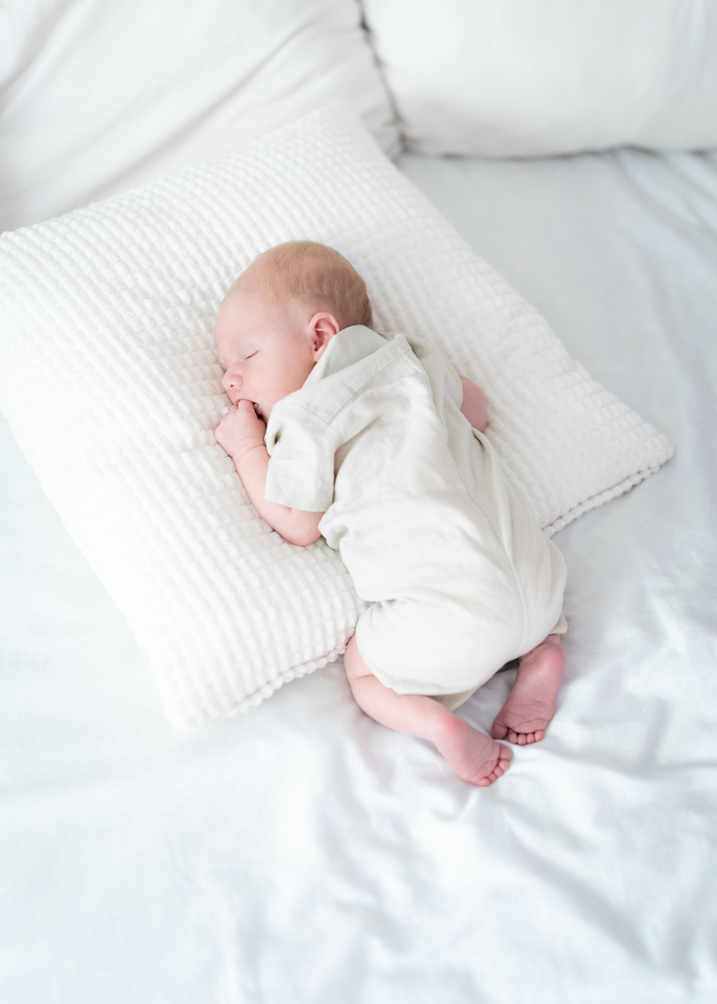 Sleeping newborn in white onesie resting on soft white pillow and bedding.