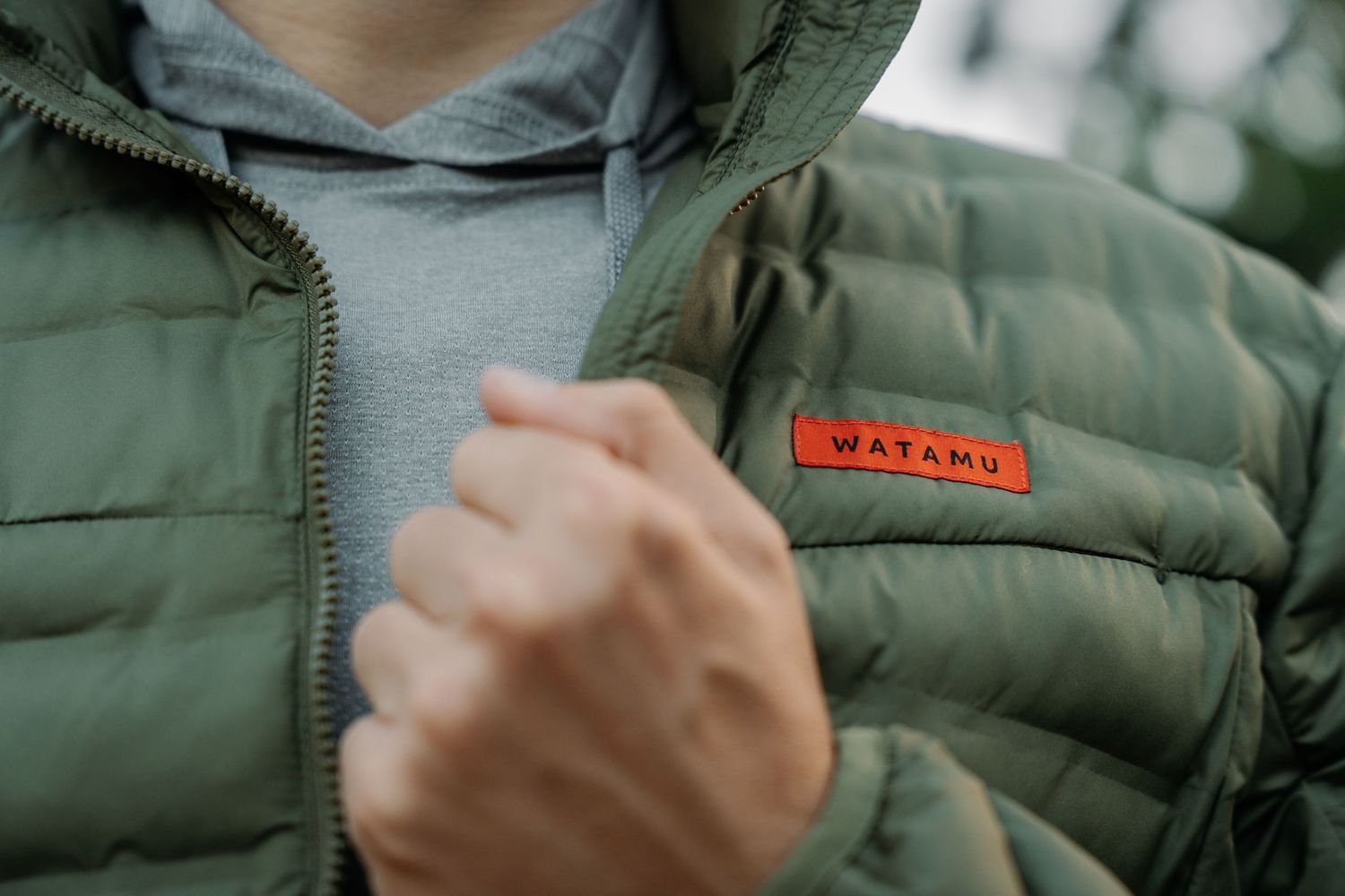 Close-up of a person wearing an olive green Watamu branded puffer jacket with a red logo patch.