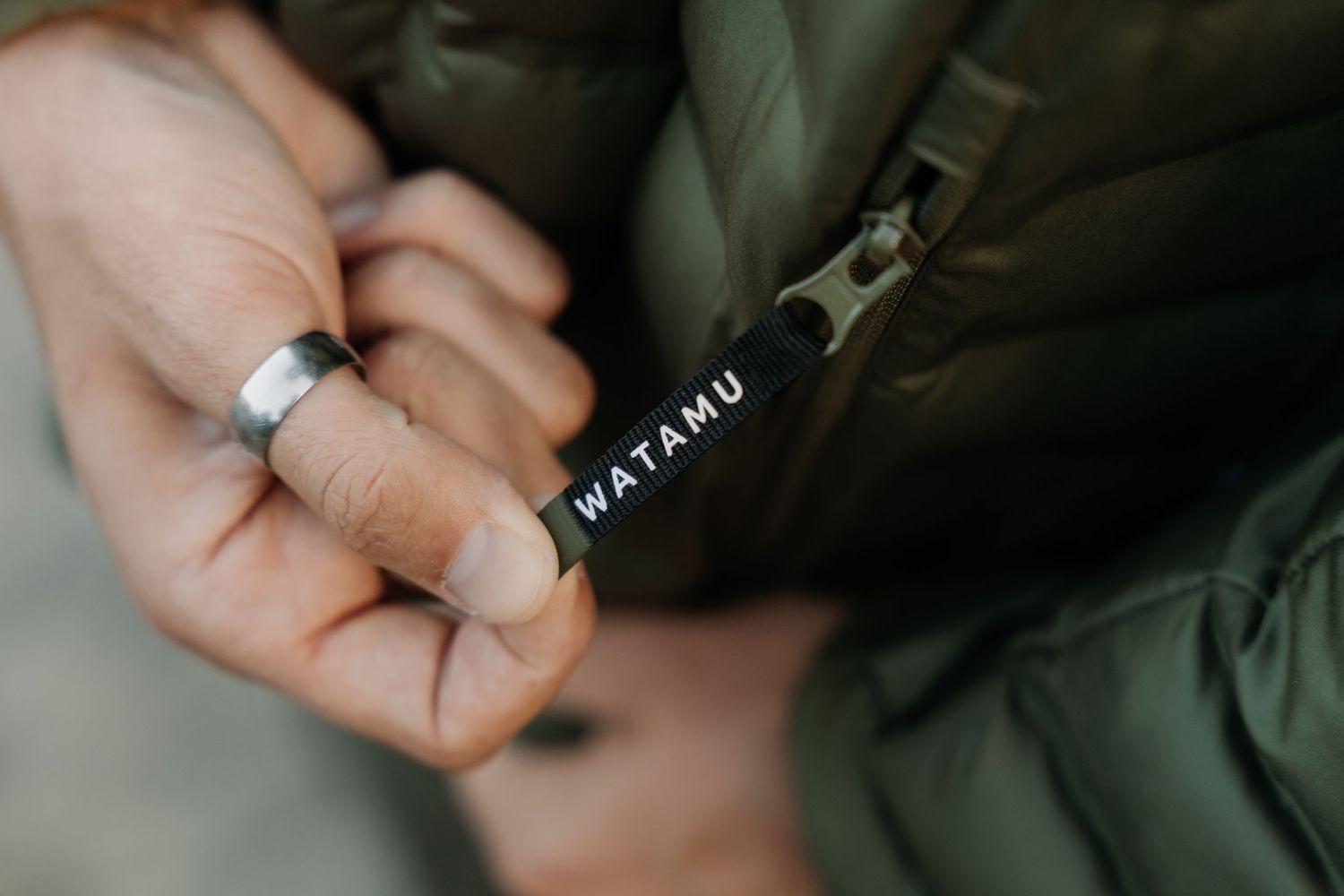 Close-up of hands holding a black WATAMU keychain tag near an olive green puffer jacket zipper.