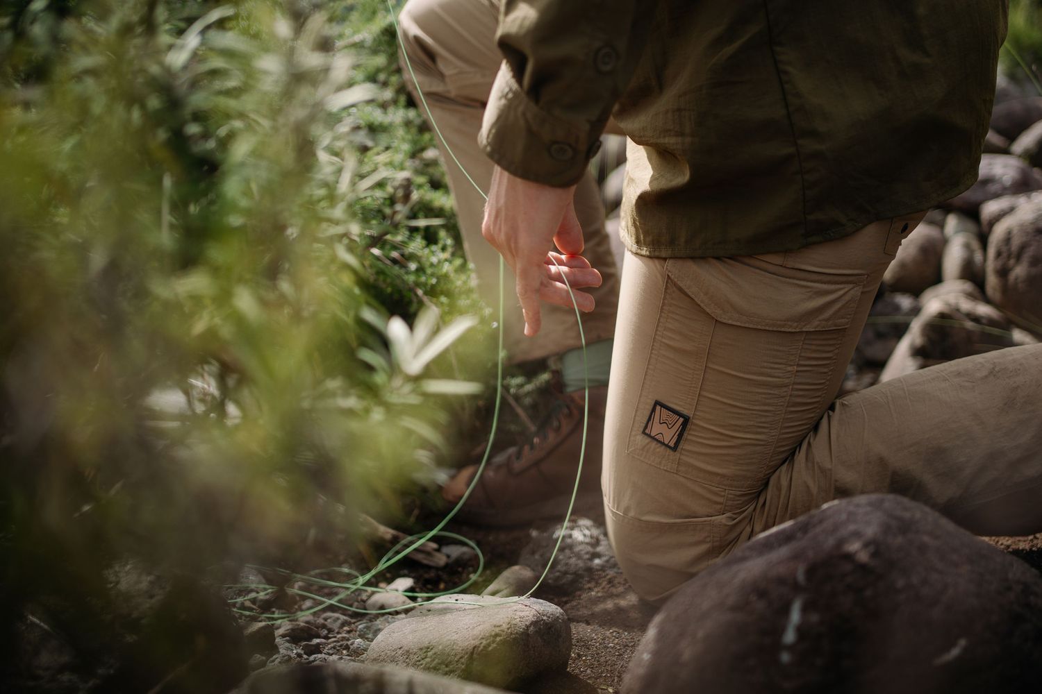 Person kneeling outdoors among rocks and plants, wearing khaki pants and olive jacket while handling equipment.