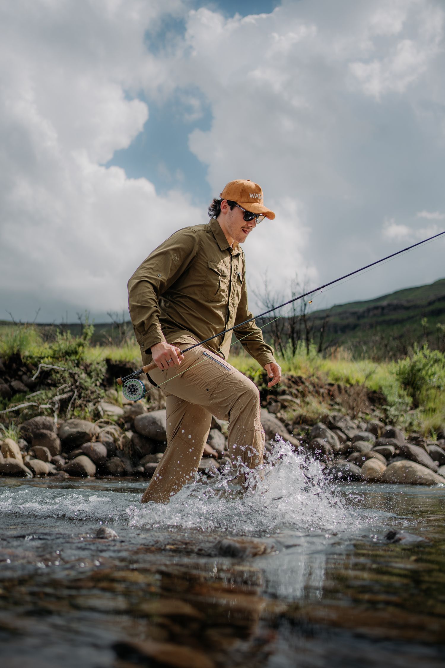 Man fly fishing in a rocky mountain stream, wearing khaki pants, olive shirt, and orange cap, splashing through clear water.