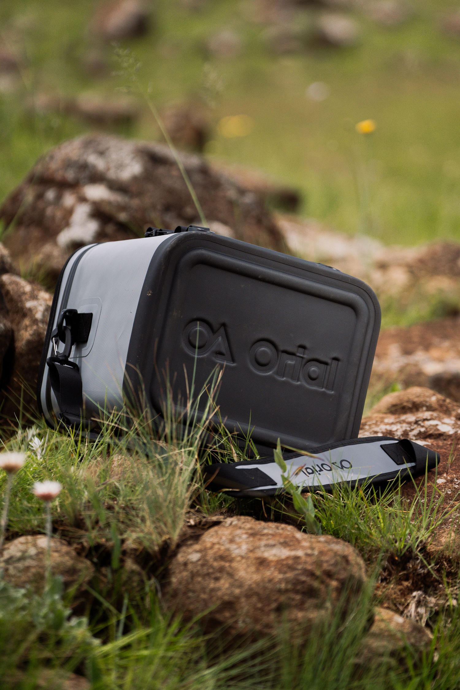 Gray Orial outdoor gear box sitting on rocky terrain with grass and wildflowers.