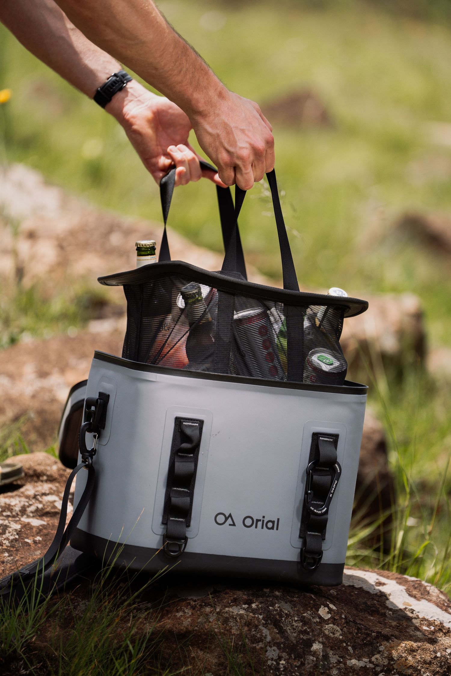 Hands lifting gear from a gray Orial outdoor container on rocky ground.