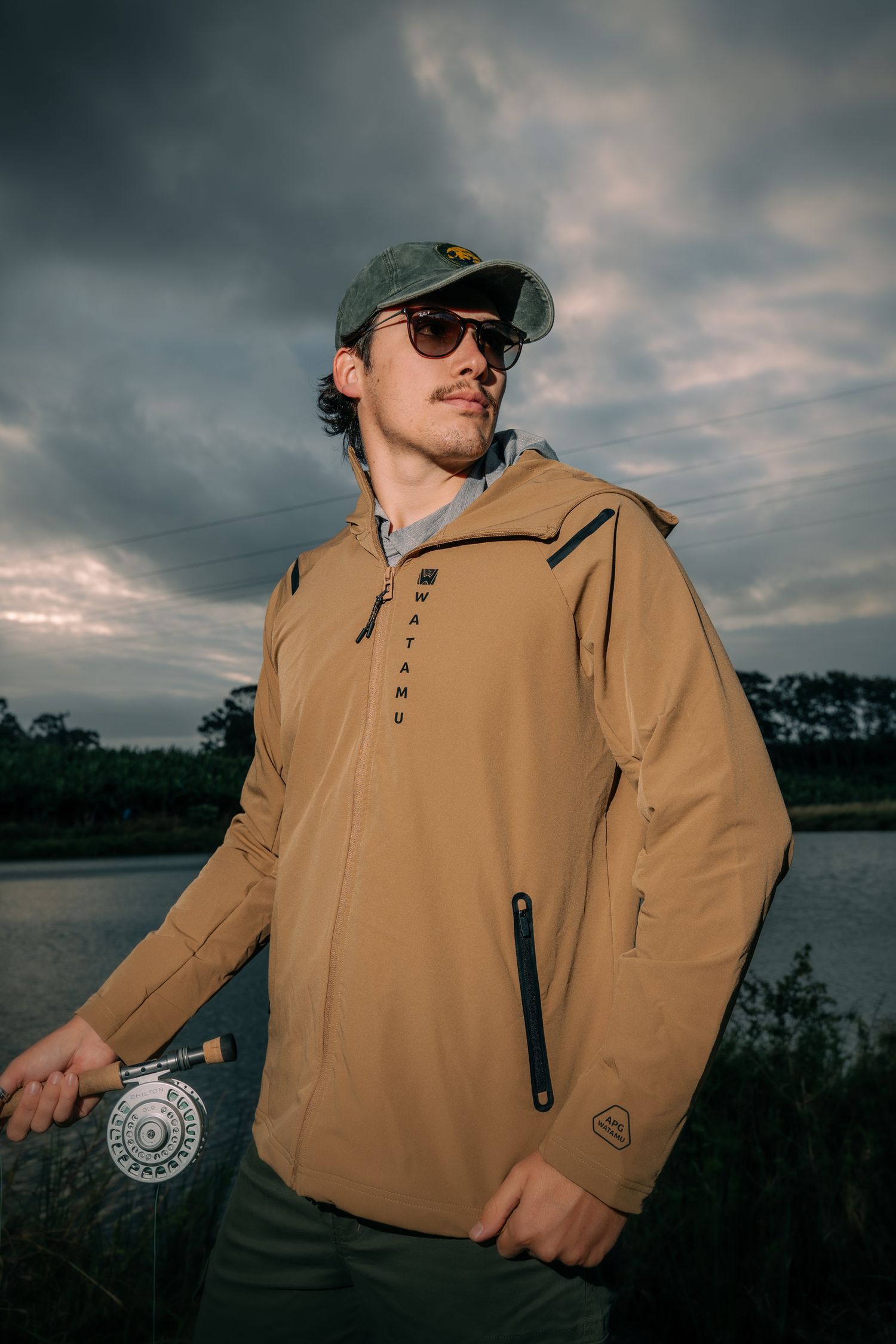 Man in tan Wtamu jacket and green cap holds fly fishing reel by a lake under dramatic cloudy skies.