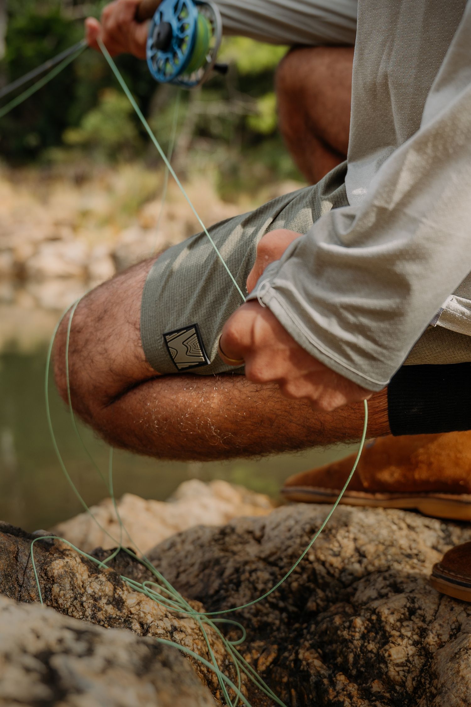 Fly fisherman tying fishing line on rocky riverbank, wearing gray shorts and white shirt with fly rod.