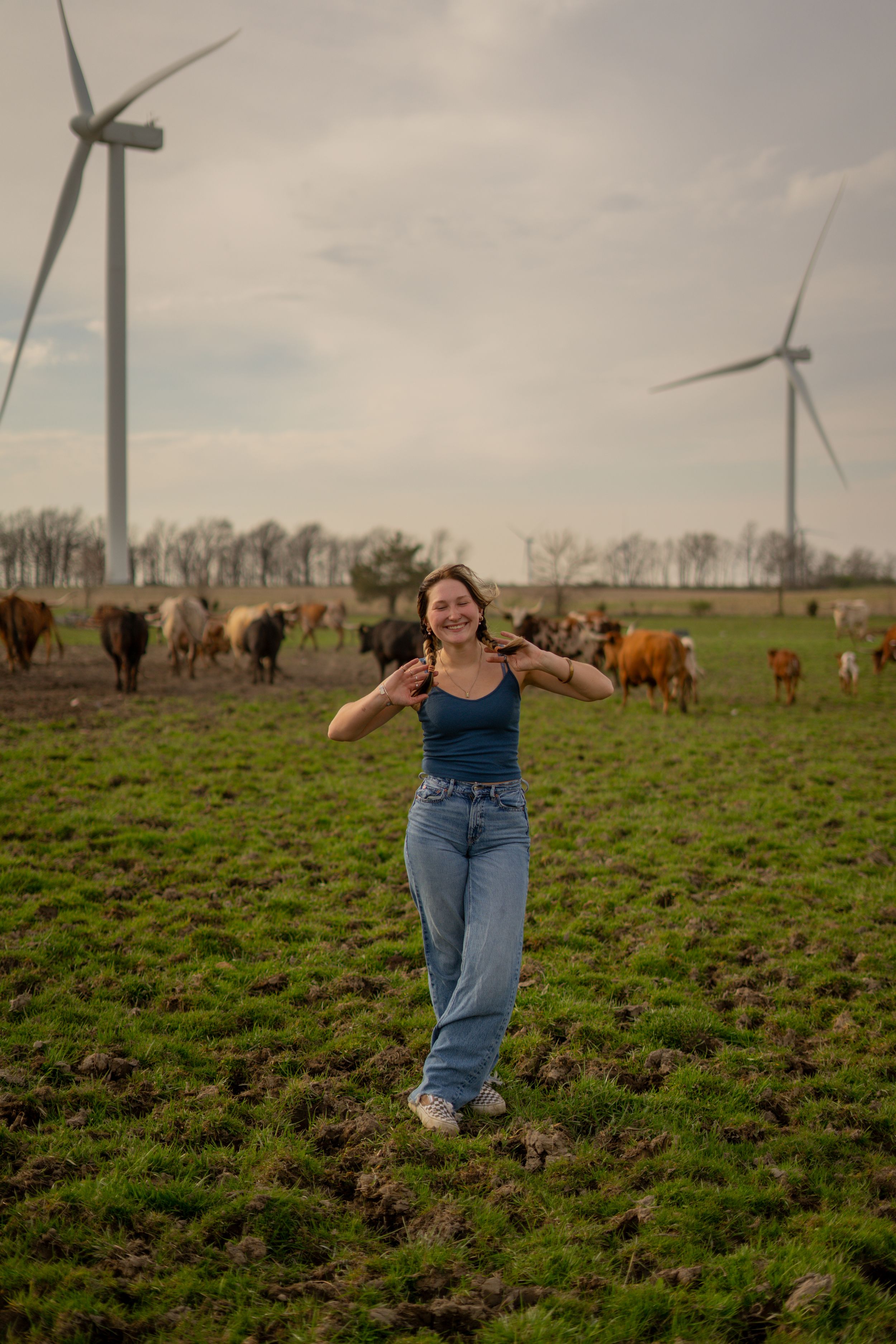 Smiling young woman in blue tank and jeans posing in a green field with cattle and wind turbines behind her.