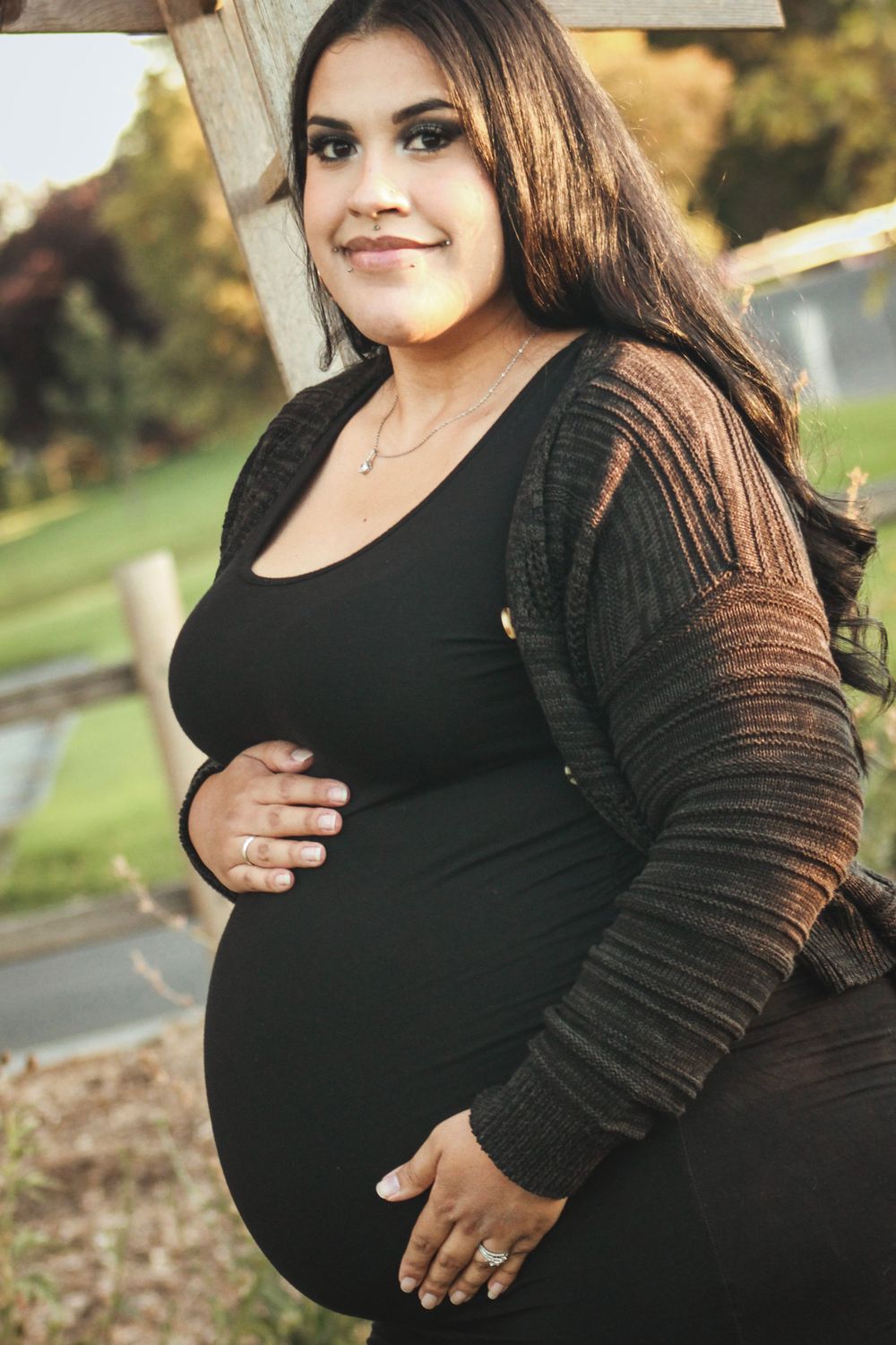Maternity portrait in black dress and cardigan against outdoor setting.