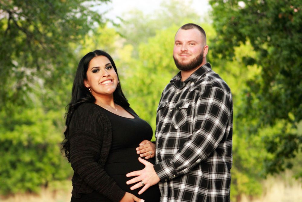 A maternity photo shoot outdoors showing an expectant couple in casual attire against a green natural background.