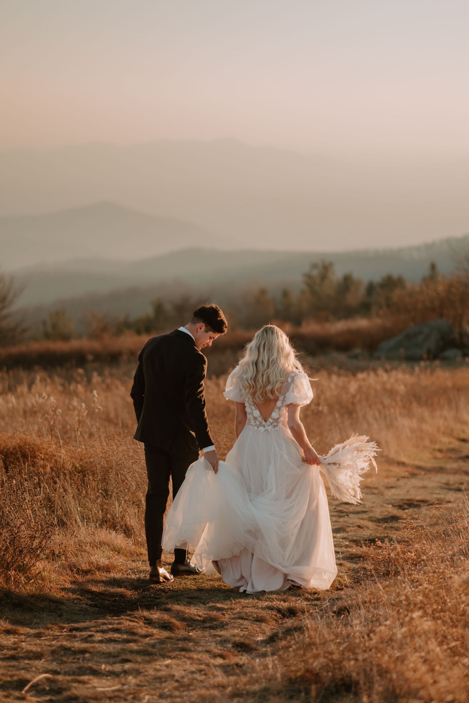 A bride and groom walk hand-in-hand through a scenic field at sunset, with mountains in the background.