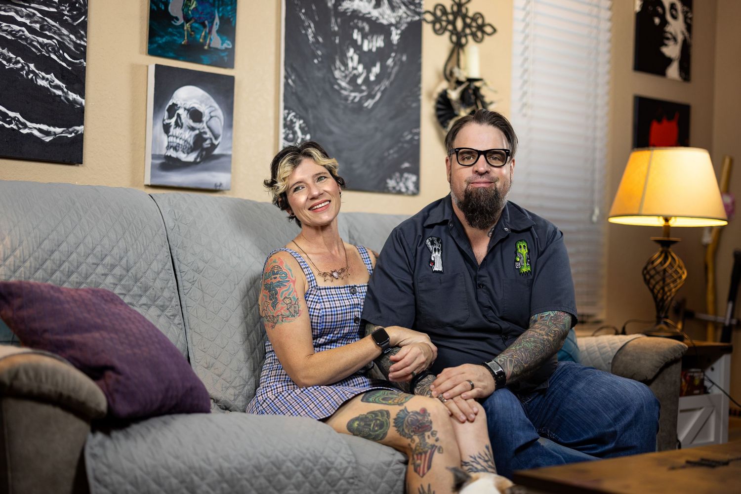 Tattooed couple smiling together on a cozy sofa surrounded by dark artwork and skull decor in a home living room.